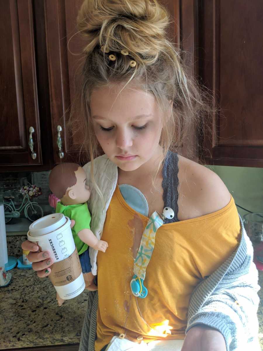 Teen dressed as "tired mom" stands in kitchen holding doll and Starbucks cup with Cheerios in her hair