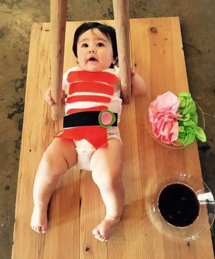 Toddler lays on wooden board dressed like sushi with two wooden sticks on either side of him like chop sticks