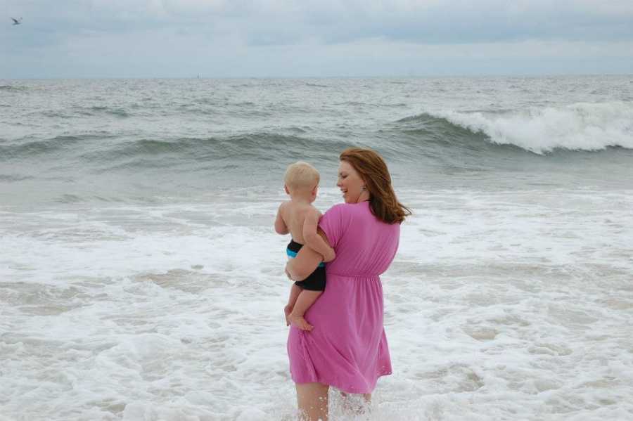 Mother stands in ocean shore holding son as wave rolls in 