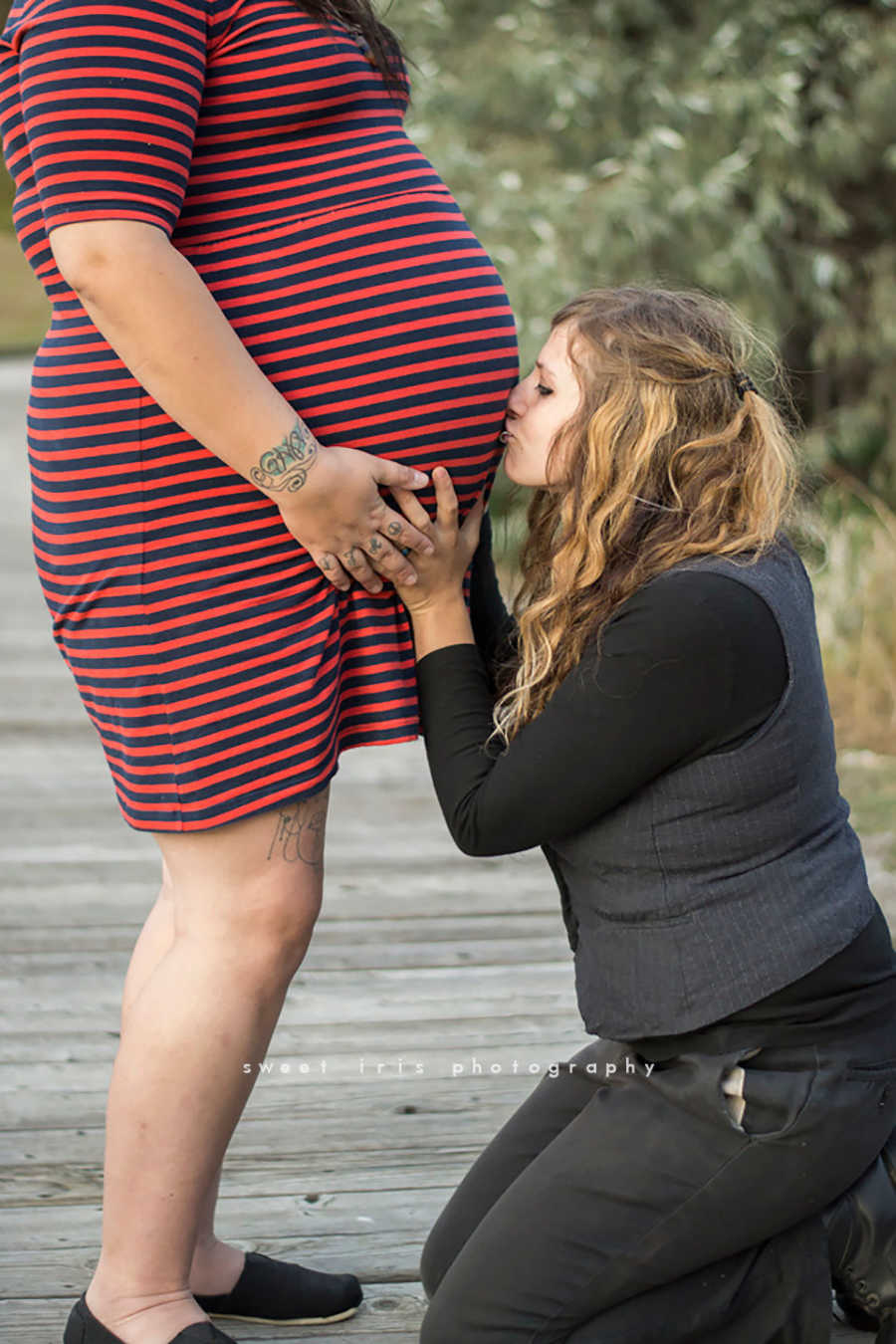 Pregnant woman stands while wife sits in front of her kissing her stomach