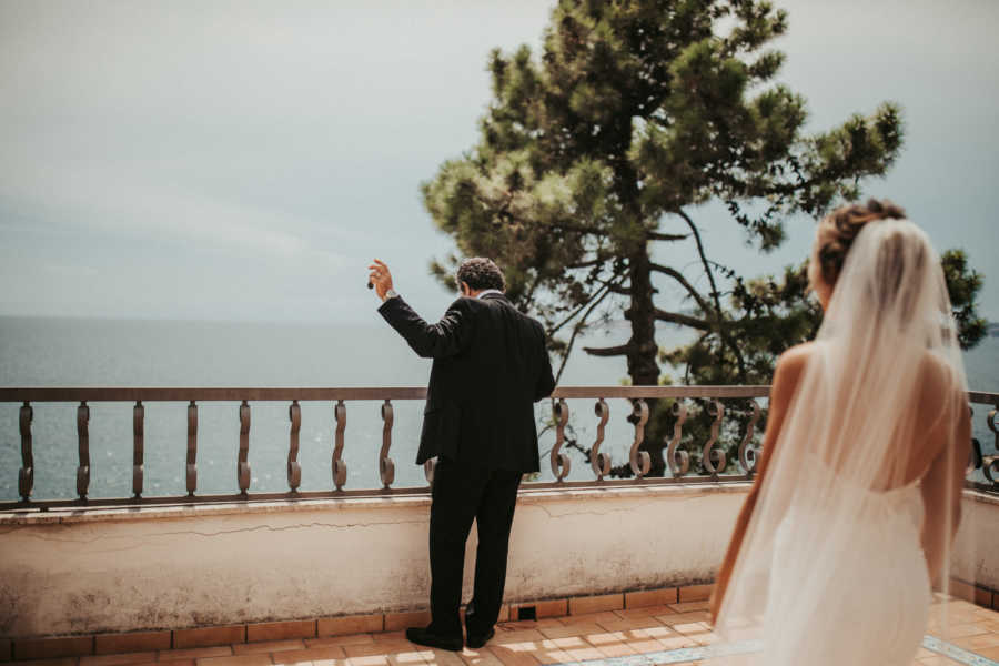 Father stands looking out at body of water while daughter stands behind him in wedding gown