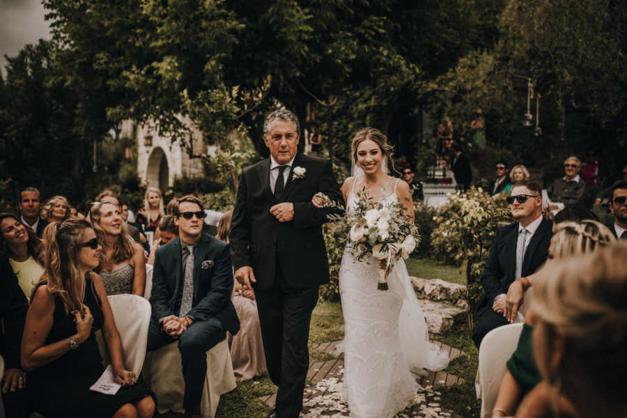 Bride smiles as she and her father walk arm in arm at outdoor wedding