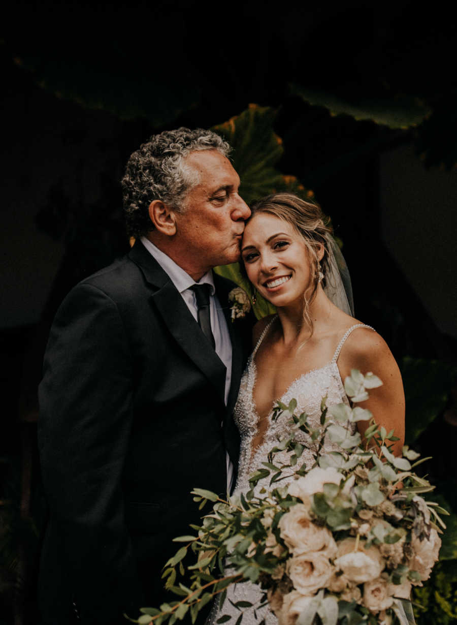 Father kisses daughter on forehead on her wedding day holding bouquet of flowers