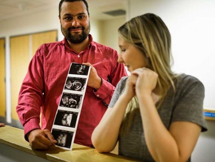 Woman stands with elbows on desk as she looks over at sonogram picture her husband holds