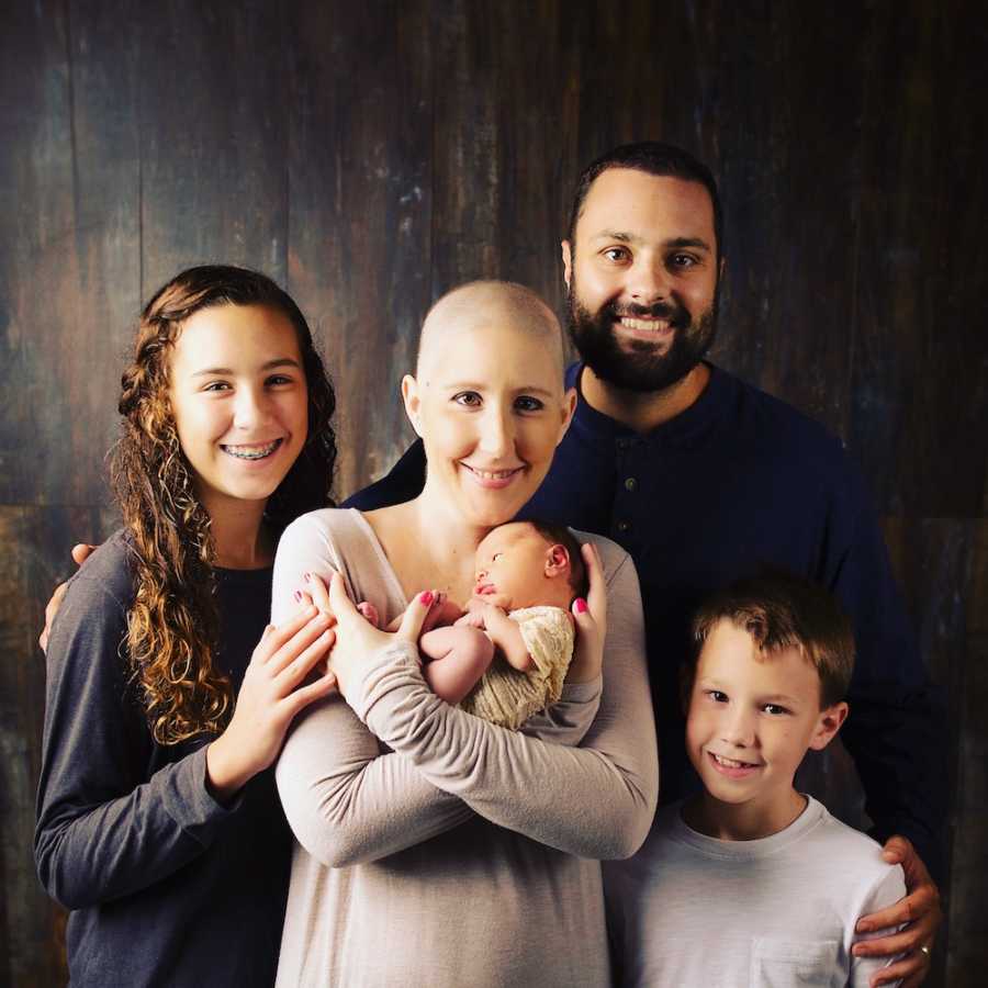 Husband and wife with breast cancer stand smiling with their two older children while wife holds newborn 