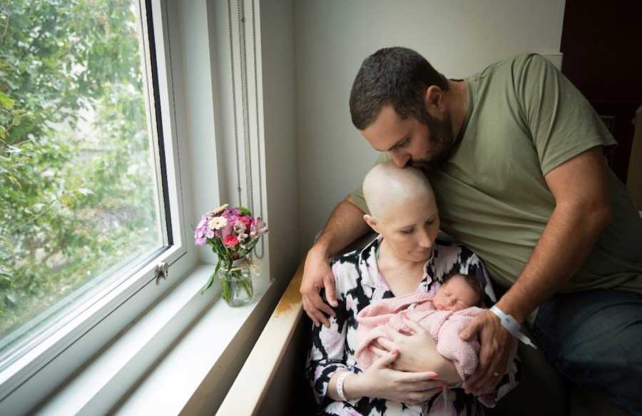 Woman with breast cancer holds newborn daughter with husband sitting beside her kissing her head