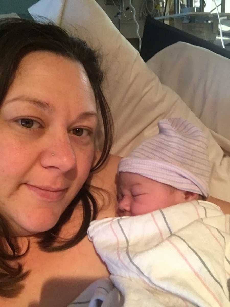 Mother smiles in selfie while lying in hospital bed with her newborn asleep in her arm