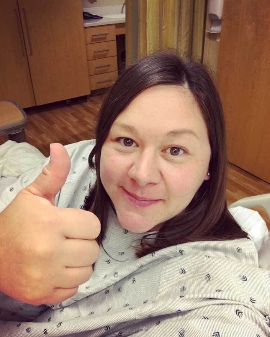 Pregnant woman smiles with thumbs up in selfie while she sits in hospital room 