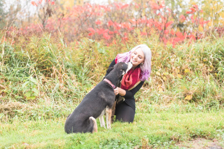 Woman sits on her knees outside while her adopted dog licks her face