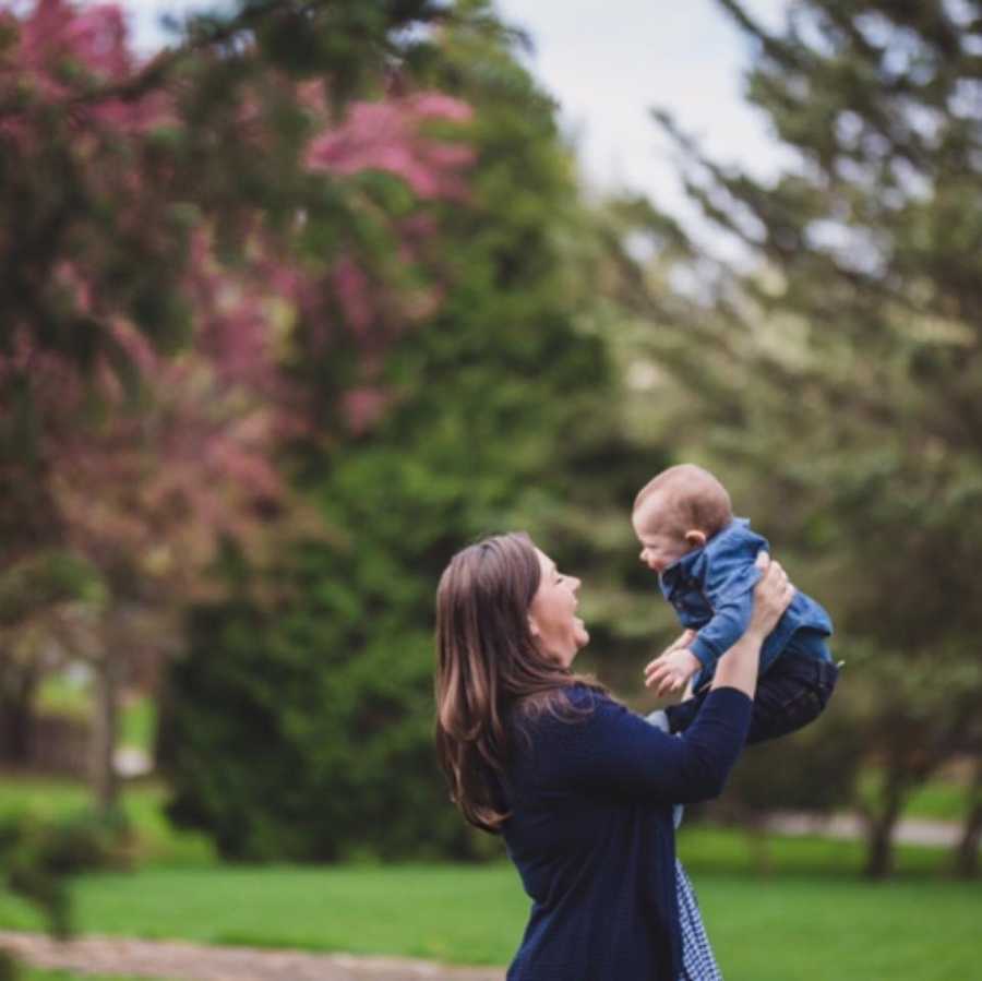 Mother stands outside smiling as she holds her baby son in the air