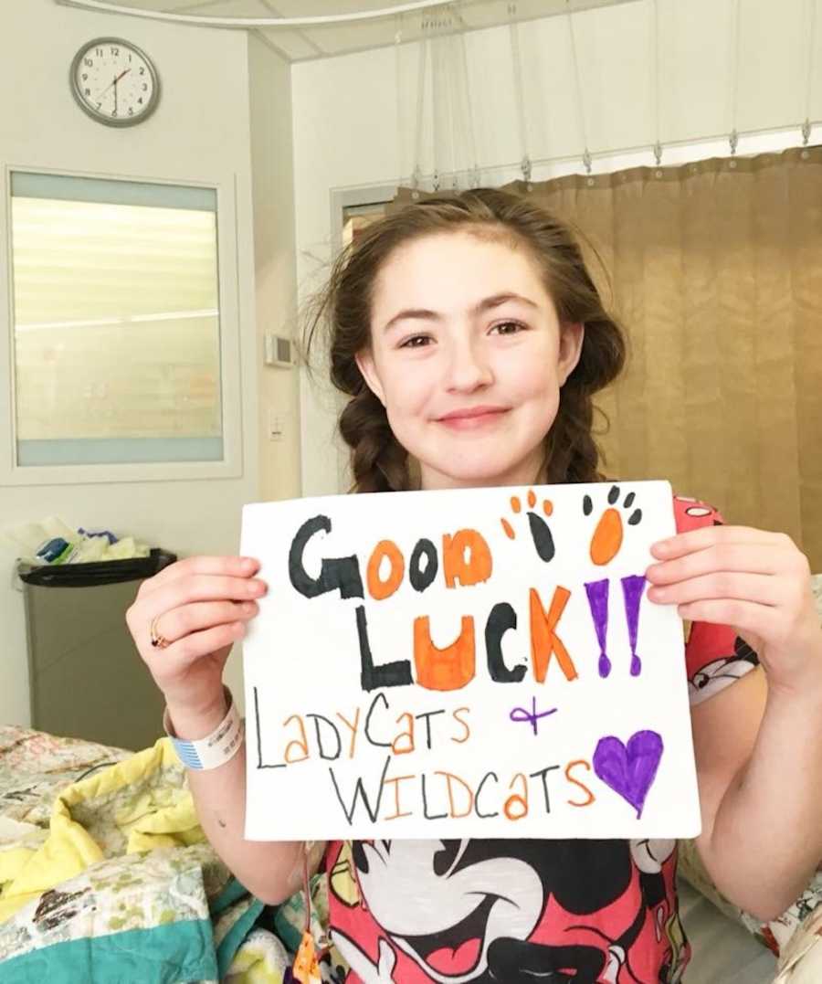 Ten year old with chronic appendicitis sits in hospital bed holding sign that says, "Good luck lady cats + wildcats 