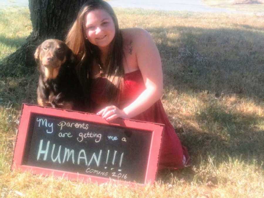 Woman sitting in grass beside dog and sign that says, "My parents are getting me a human"