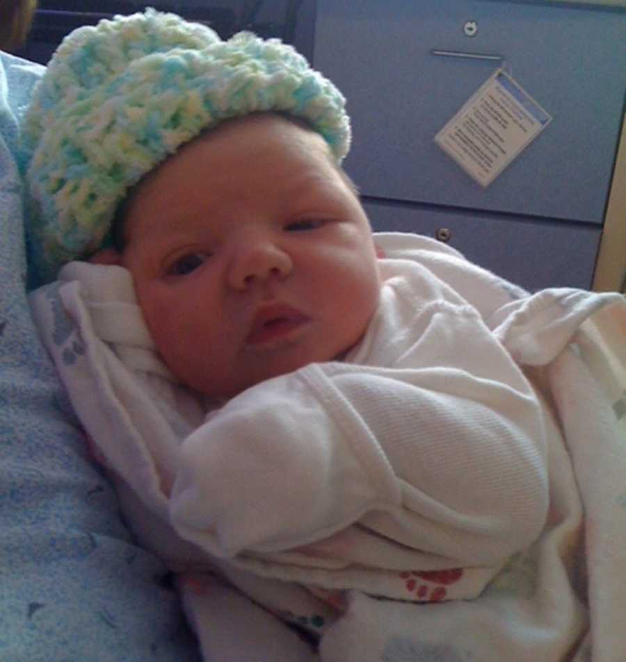 Newborn baby with CMTC in white onesie and blue, white and green knit hat