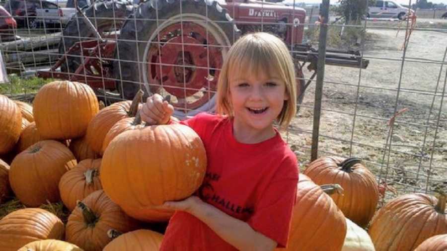 Young girl smiles as she stands holding pumpkin in pumpkin patch