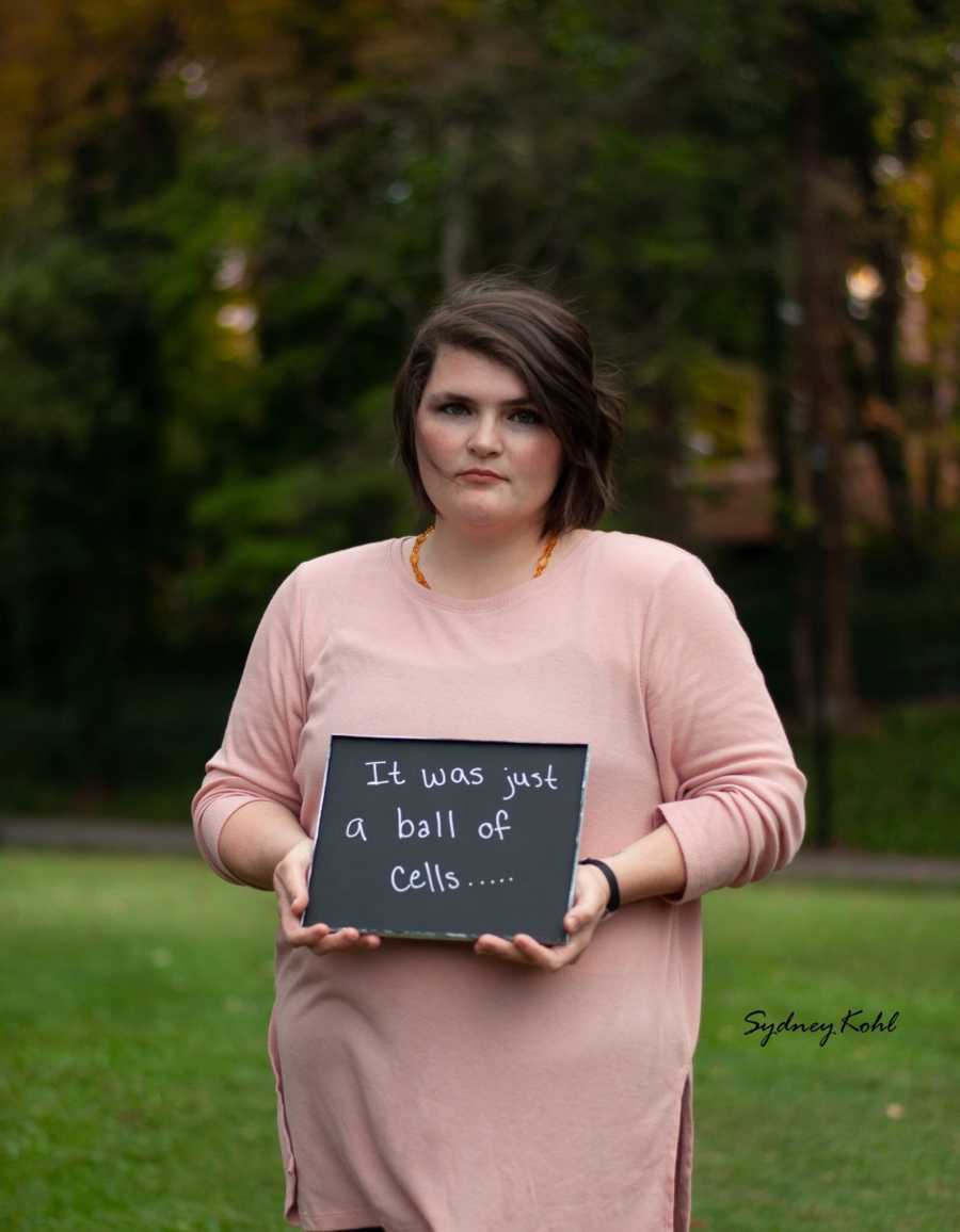 Woman who went through child loss holds sign that says, "It was just a ball of cells"