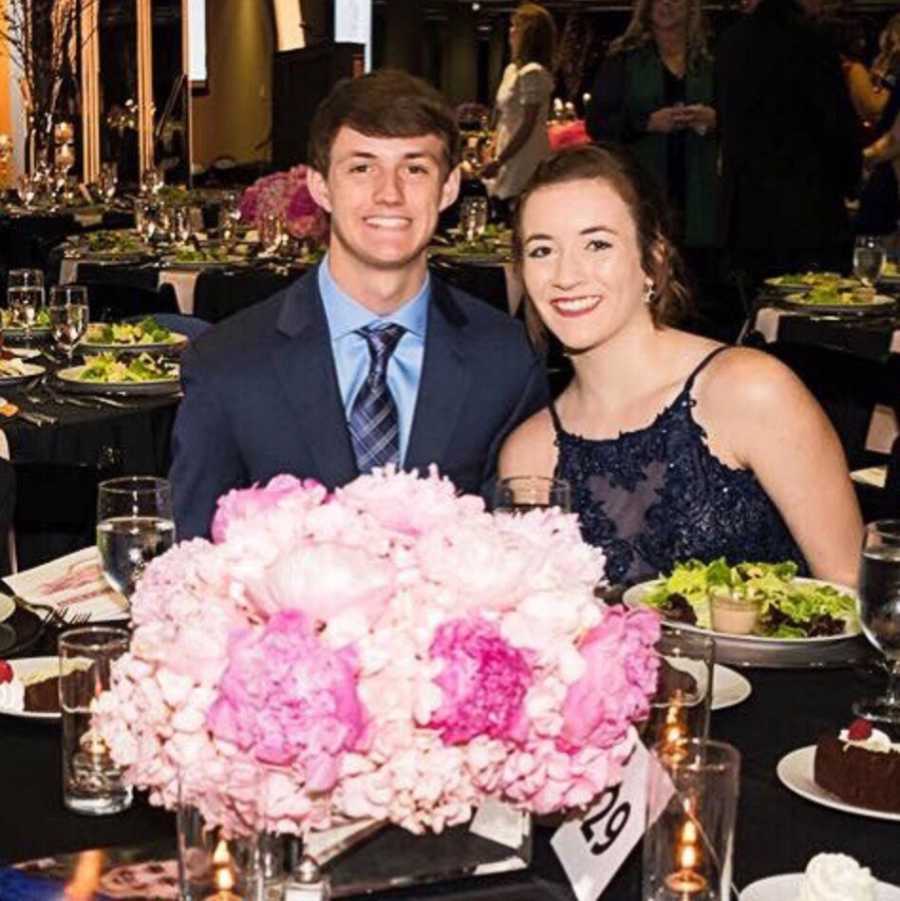 Teen boyfriend and girlfriend sit at wedding reception smiling 