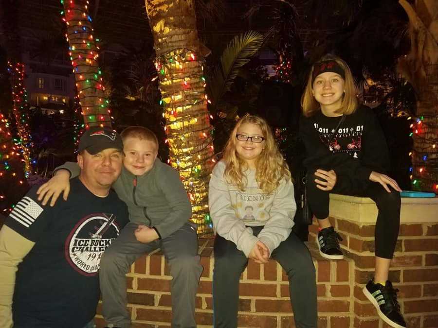 Father smiles with arm around son who sits beside his wife's two daughters on brick wall