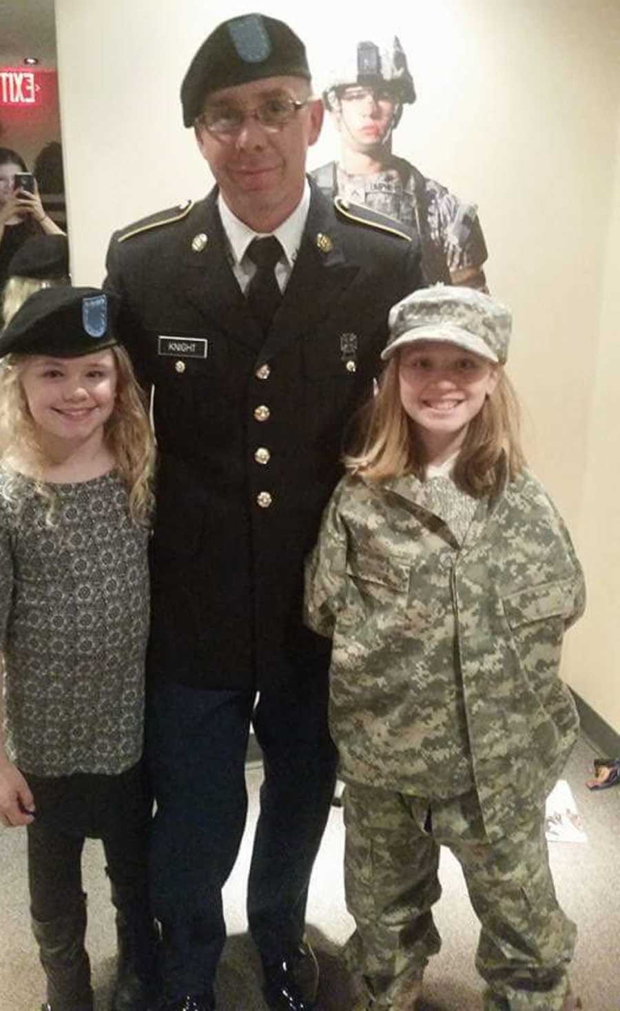 Army man stands in uniform with his arms around his wife's two daughters