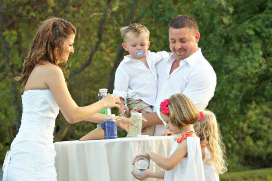 Bride stands beside her two daughters at high top table while groom stands holding his son