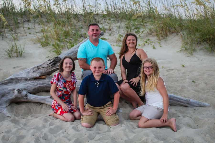 Husband sits on branch on beach beside wife, son, and his two adopted daughters