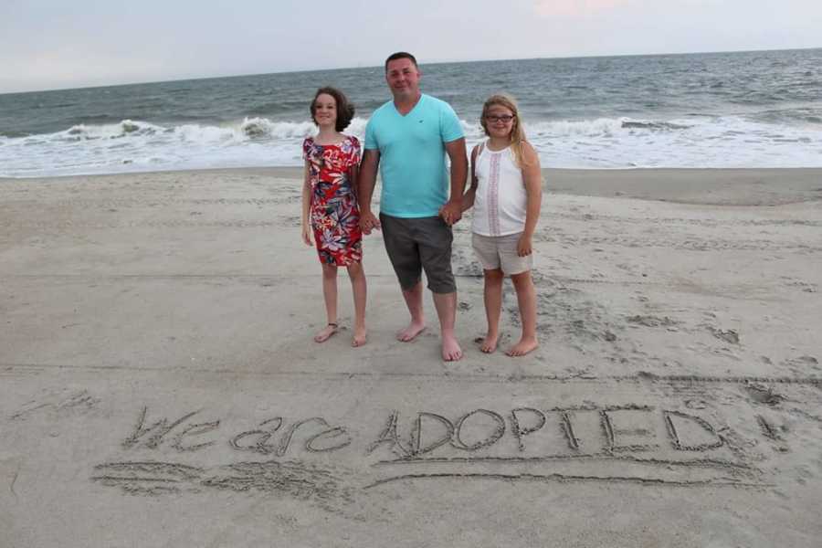 Man holds wife's daughters hands on beach and written in the sand says, "we are adopted"