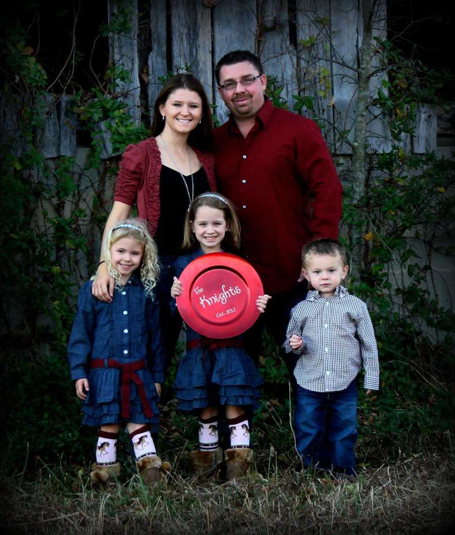 Husband smiles with his young son beside his wife and her two daughters, one of which is holding sign saying, "The Knights"