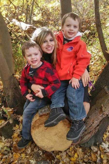 Single mother sits on tree stump in woods with her twin boys on her lap