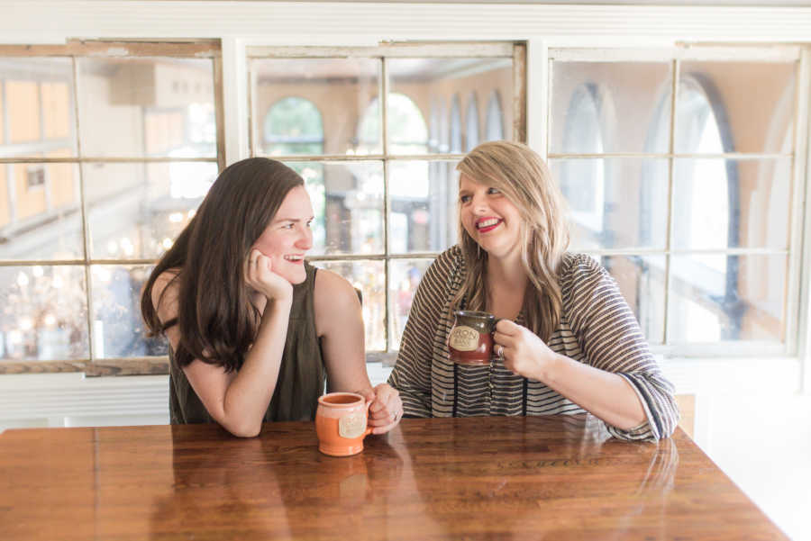 Woman sits at table smiling at friend as they hold coffee mugs