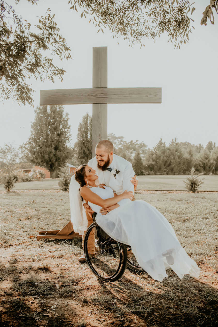 Groom drips paralyzed bride in wheelchair in front of large cross outside