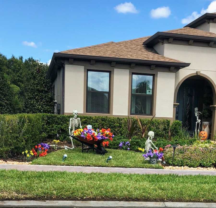 Skeleton in yard holding wheelbarrow full of flowers while the other skeleton sits on ground planting them