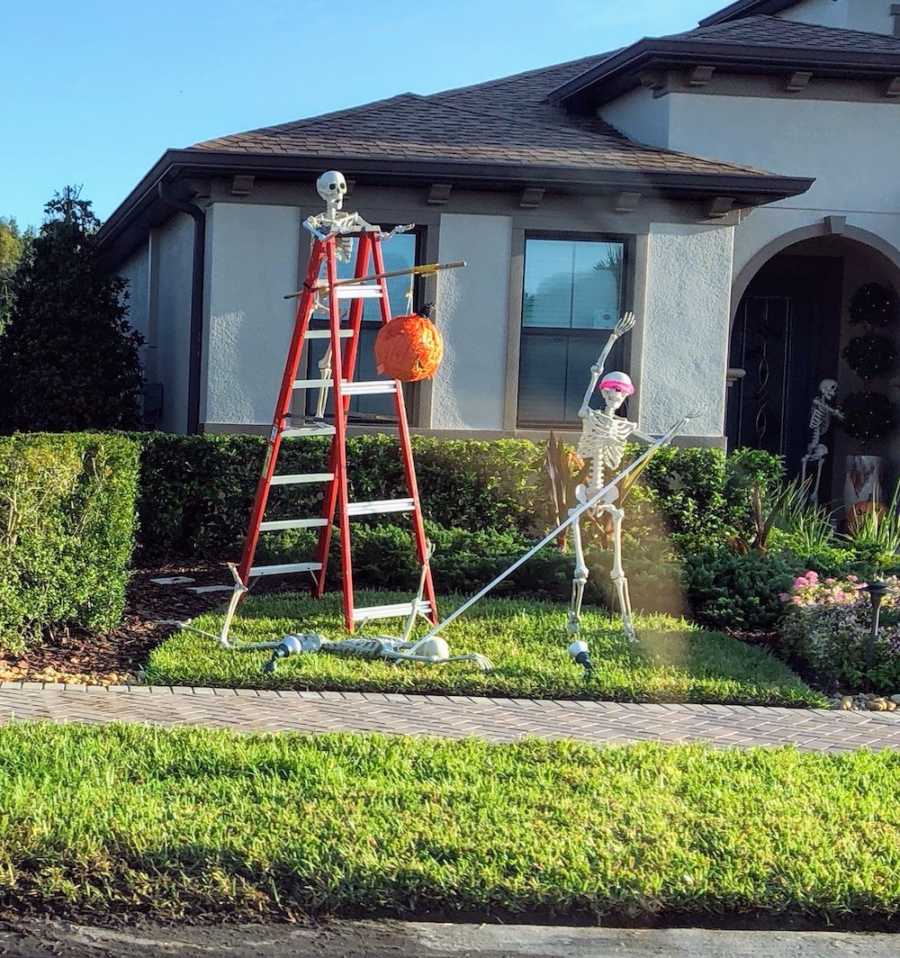 Skeleton stands on later holding orange pinata while another skeleton who is blindfolded hits skeleton who is laying on ground
