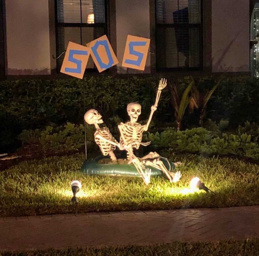 Skeletons sitting in yard on inflatable raft if "SOS" sign behind them