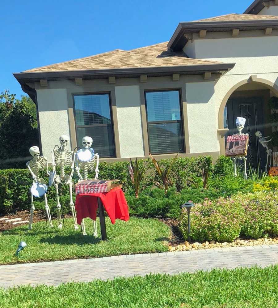 Three skeletons stand in yard holding paper plates while one stands by table opening pizza box