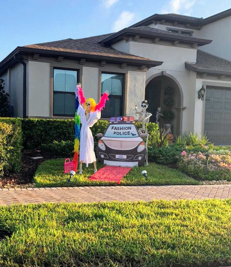 Skeleton wearing robe and boa stands with hands in air in front of "Fashion Police" cop car and skeleton with water gun