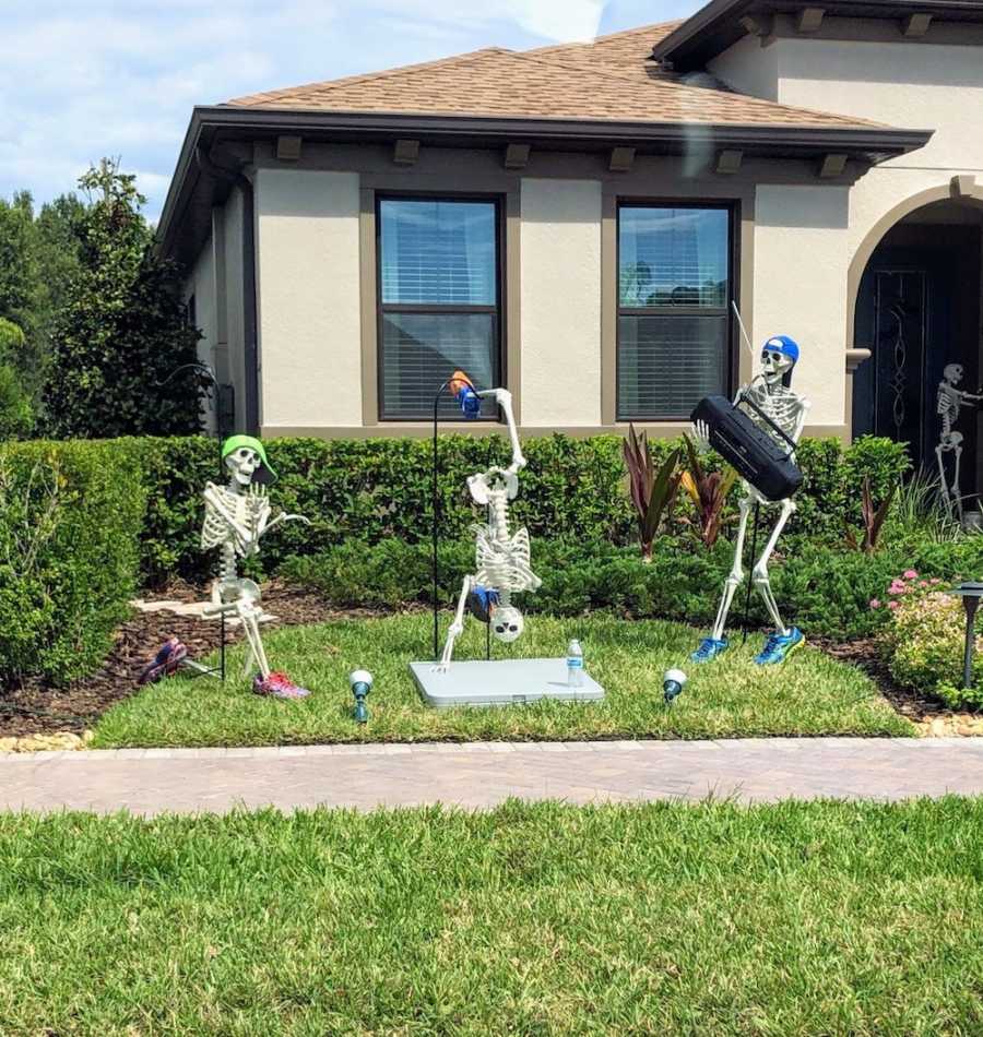 Skeleton stands in yard holding boom box while another skeleton break dances and other sits watching