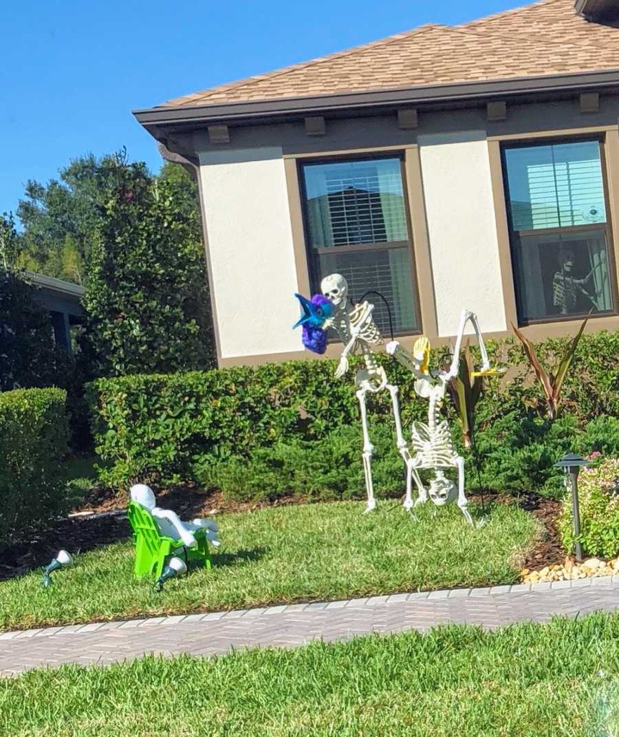 Small skeleton sits in chair in yard watching one hold up bird head and other doing handstand