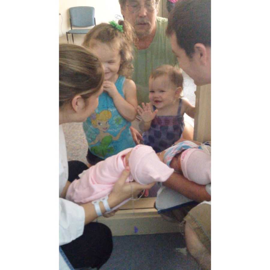Mother and father hold twin newborns through window so that their two other daughters can see