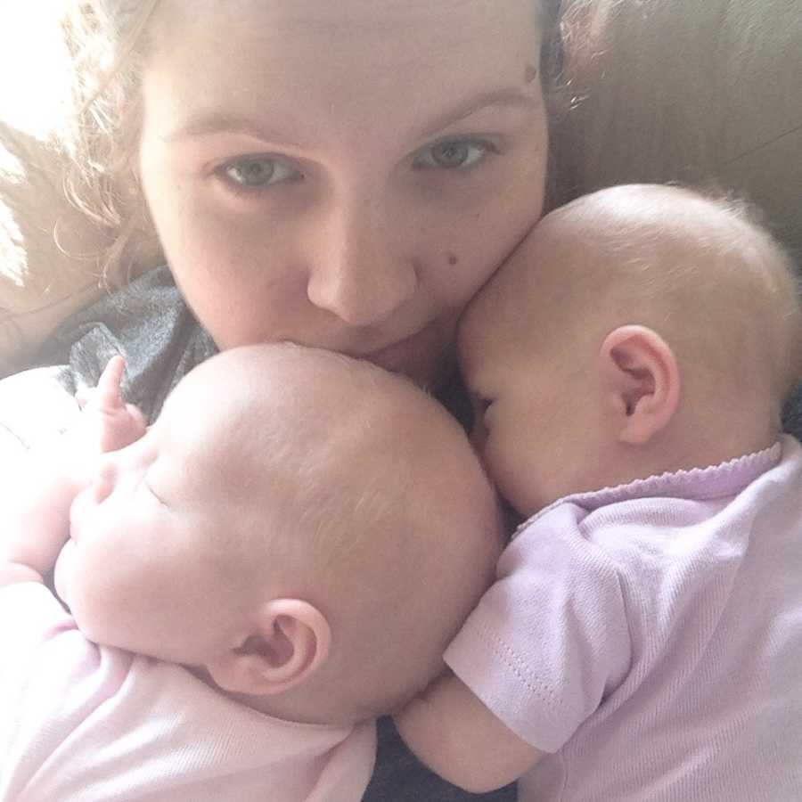 Mother smiles in selfie with twin daughters asleep on her chest