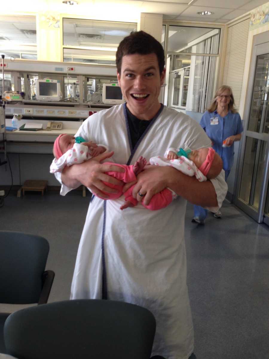 Father stands smiling holding twin daughters in his arms