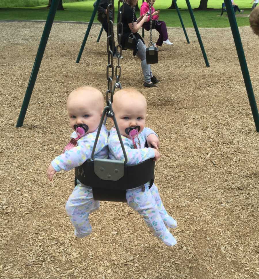 Baby twin girls sit back to back on baby swing on playground