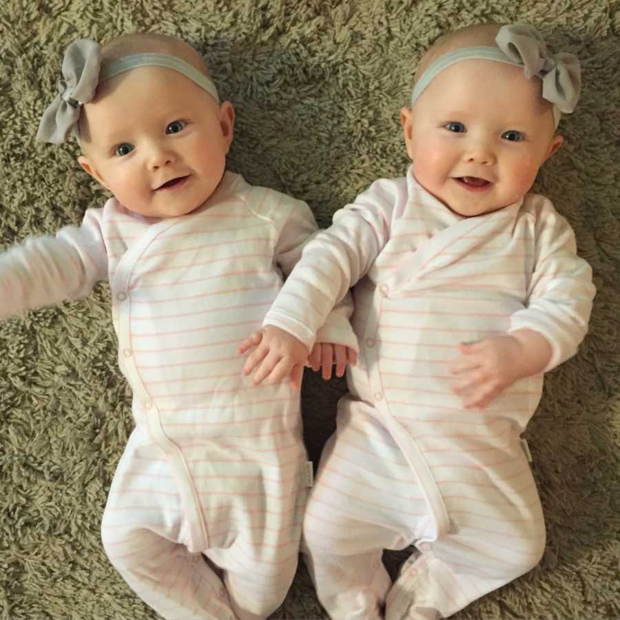 Baby twin girls laying on floor wearing pink and white onesie with gray headband