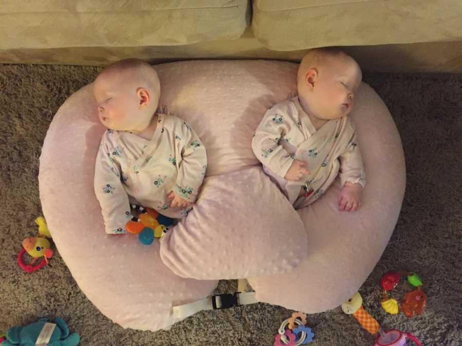Baby twin girls lay asleep on pink pillow in matching onesie with toys surrounding them