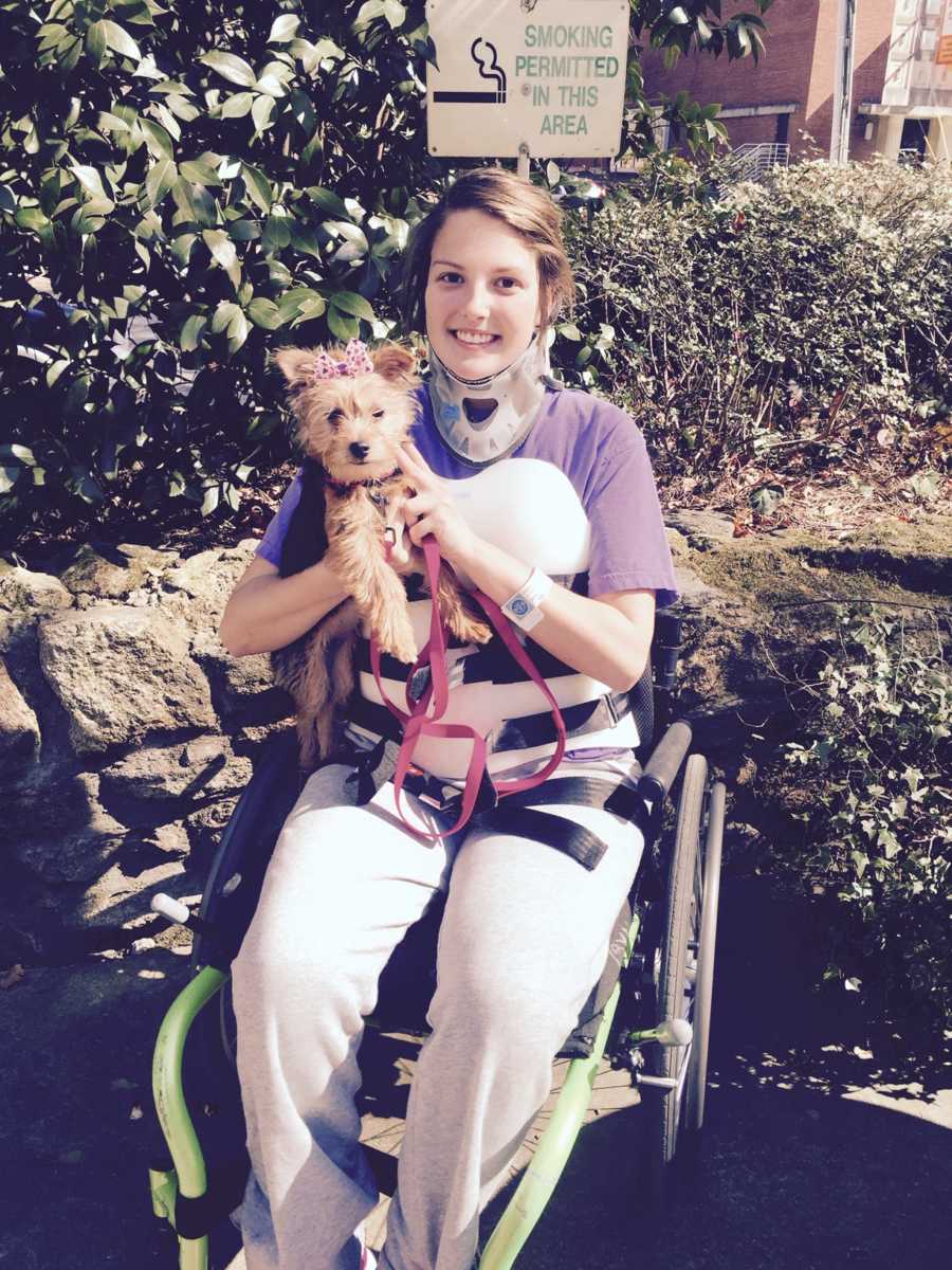 Woman sits in wheelchair wearing back brace while holding little dog