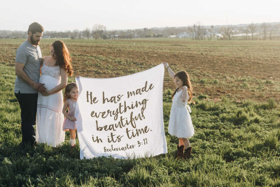 Pregnant woman stands in field with her husband holding her stomach beside daughters who hold flag