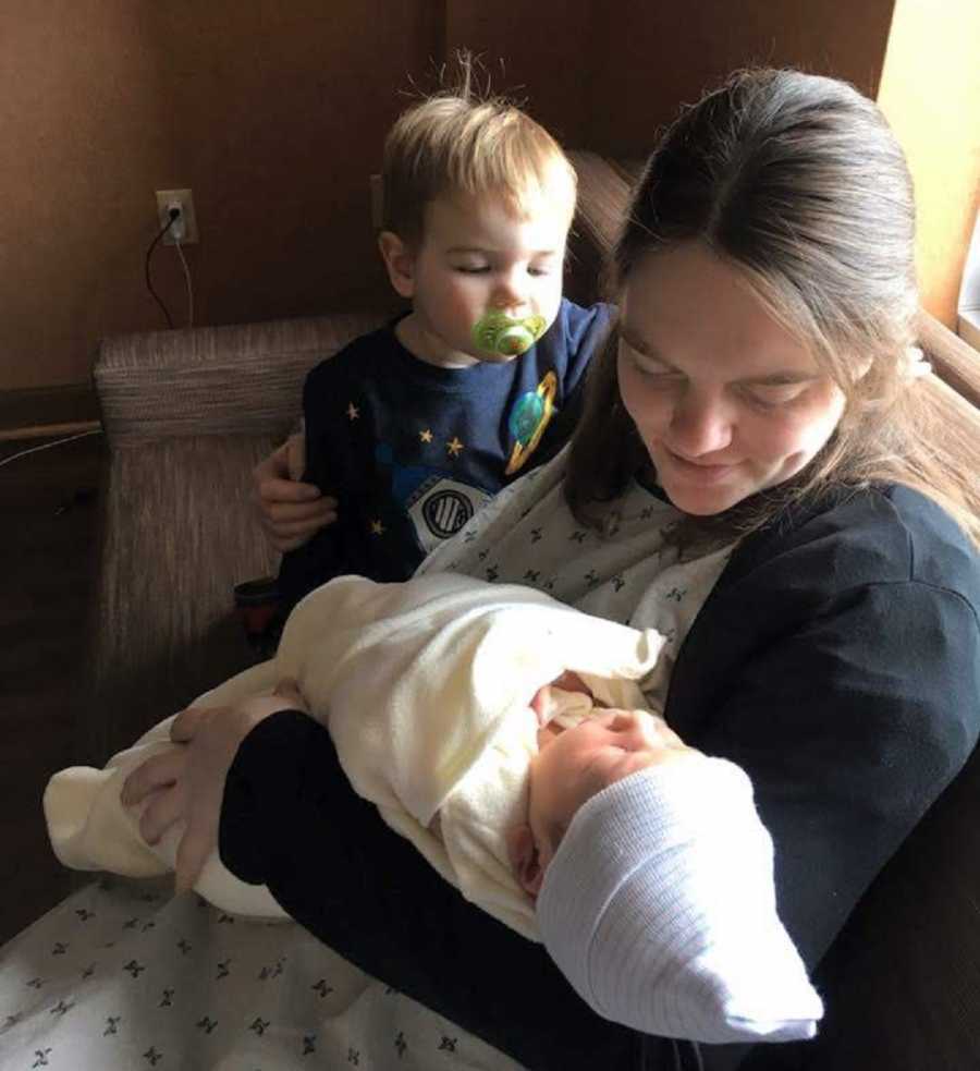 Mother sits on couch looking down at newborn in her arm with firstborn looking on beside her