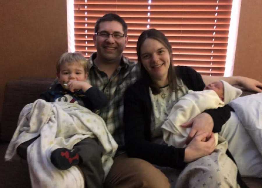 Husband and wife sit on couch with their firstborn while wife holds newborn in her arms