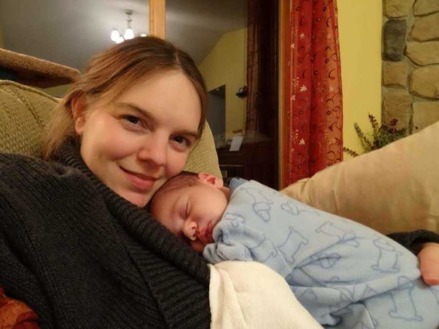 Mother smiles in selfie whole sitting in chair at home with newborn son asleep on her cher