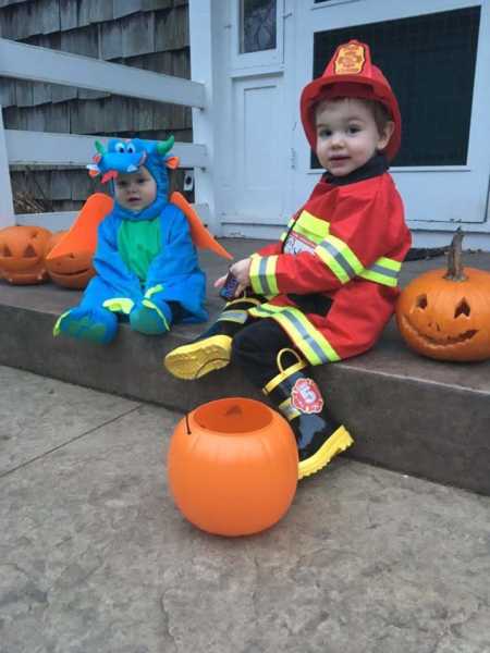 Two young boys in fireman and dinosaur costume sit on ledge by front door beside jack-o-lanterns