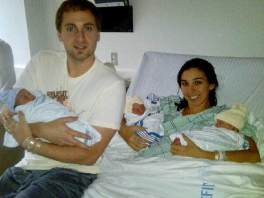 Woman lays in hospital bed with two of her triplets in her arms beside husband who holds third 