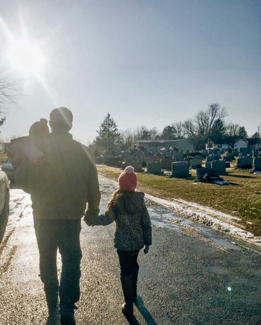 Father holds one daughter and holds hand of other as they walk in cemetery 